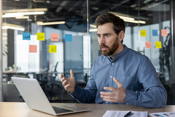 A serious young male businessman sits in the office at his desk and looks shocked at the laptop...