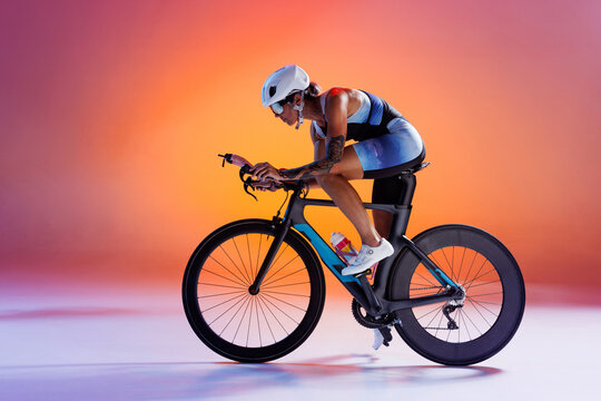 Cyclist in colorful sportswear and white helmet riding road bike against gradient orange and pink background in neon light. Concept of sport, competition, tournament, championship, speed, endurance