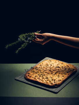 a hand holding a sprig of rosemary over a bread on a green surface with a black background