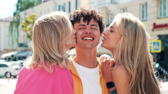 Three stylish friends posing in the street. Fashion man and two cute female dressed in casual summer clothes. Smiling models having fun. Cheerful women and guy outdoors. They kiss him in cheek