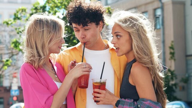 Young three stylish friends posing in the street. Fashion man and two cute female dressed in casual summer clothes. Smiling models having fun. Cheerful women and guy outdoors, Hold and drink lemonade