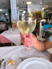 Hand of a man, sitting at a table, holding a glass of white wine, against a restaurant background. Hand of a man with a glass of white wine toasting. Glass of white wine held in hand by a man.