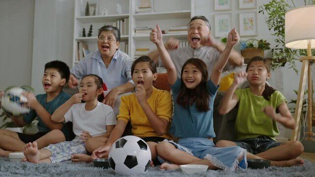 asian family sitting on couch at home watching live broadcasting of soccer game together celebrating a goal,Grandfather and grandmother watch a football match streaming TV with their grandchildren.