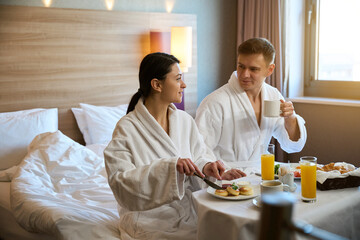 Newlyweds in bathrobes having breakfast together in hotel room
