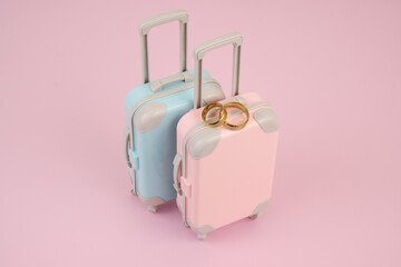 Pink and blue suitcases with two golden wedding rings on pink background. 