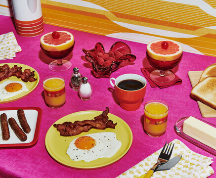 Fototapeta Breakfast table of eggs, bacon, fruit, coffee, and orange juice in a vintage setting