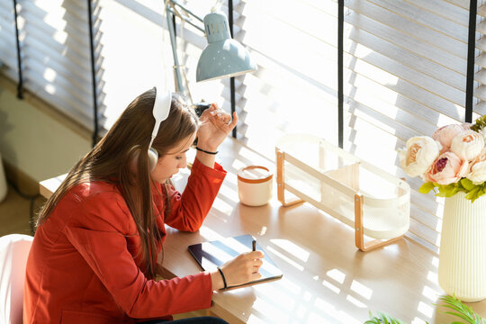 young woman wearing a red shirt sits listening to music and working in the living room next to her window during the morning sunlight. in a comfortable atmosphere happily