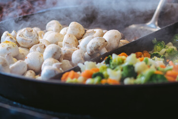 Mushrooms and other chopped fresh vegetables are fried in a hot frying pan outside. Healthy vegetarian food. Fair of street food. Space for text