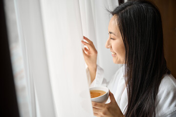 Attractive Asian woman opening window with white tulle and looking outside