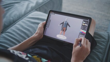 Focused teenager draws clothes for game character using 3d modeling program. Boy with digital pencil and tablet in hands sits in room, creates 3d models. Game design and development concept. Close up.