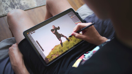 Boy with digital stylus and tablet in hands sits in cozy room and draws 3d model. Teenager creates new skin using 3d modeling program. Young game designer remote studying. Game development. Close up.