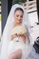 A bride is holding a bouquet of white flowers in her lap. She is wearing a white dress and a veil....