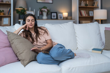 Obraz premium Simple living. Young beautiful relaxed woman sitting alone at home on sofa, listening podcast or music on her wireless headphones connected to her smart phone. Freedom concept and weekend activities.