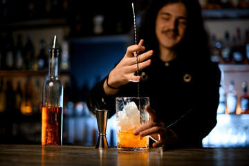 Smiling bartender with a metal details for making cocktails and glass of whiskey with ice