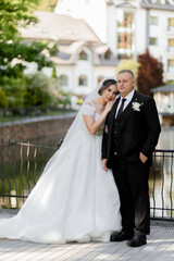 Fototapeta premium A bride and groom pose for a picture on a bridge. The bride is wearing a white dress and the groom is wearing a black suit