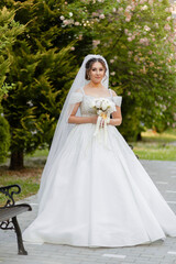 A woman in a white wedding dress is holding a bouquet of flowers. She is standing in a park, surrounded by trees and benches. Concept of celebration and happiness