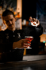 Slim cocktail maker in black shirt stands with glass with whiskey or cognac