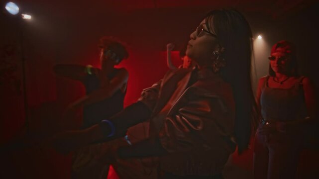 Handheld lowkey shot of group of young energetic ravers in trendy y2k outfits dancing to electronic music and enjoying night in club with red neon lights and repeated flashes