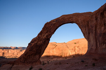 Wide shot of Corona Arch in Moab, Utah, showcasing its stunning sandstone formation in shade at sunrise. The natural arch stands prominently, with the background landscape bathed in sunlight - USA