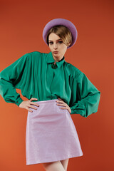 A young woman in a green blouse and lavender skirt poses against a bright orange backdrop.