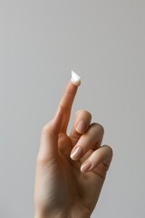 Detailed close-up of a hand with well-manicured nails holding a dab of cream on the fingertip against a neutral background.
