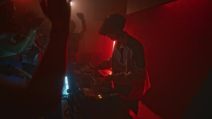 Side shot of cool dj in tracksuit playing electronic dance music on audio mixer to raving crowd during retro party in red neon lit nightclub
