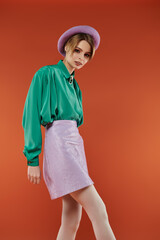 A woman in a green shirt and purple skirt stands confidently in a studio setting.