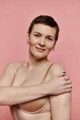 A woman with short hair, wearing a bra with a pink ribbon, smiles confidently against a pink background.