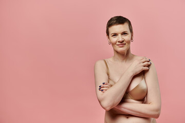 A woman with short hair and bra smiles confidently against a pink background.