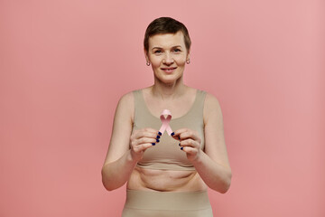 A woman with short hair, wearing a bra and holding a pink ribbon, smiles at the camera.