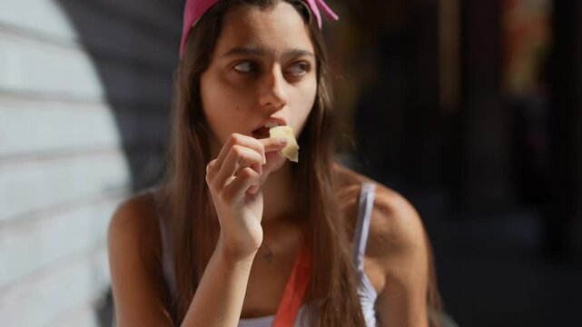 A young woman is sitting outdoors, enjoying a snack in her casual summer attire