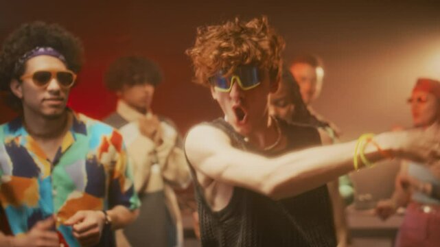 Handheld shot of energetic curly-haired guy in mirrored techno rave sunglasses and mesh tank top rocking out and making powerful movements while dancing to camera at retro party in nightclub