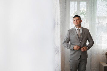 A man in a suit is standing in front of a window, adjusting his tie. Concept of formality and professionalism, as the man is dressed in a suit and tie