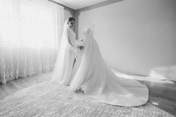 A bride is getting ready for her wedding and is adjusting her dress. The bride is standing in front of a white dress and a white curtain