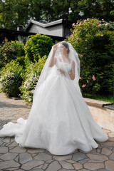 A woman in a white wedding dress is standing in front of a house with a green hedge. She is wearing a veil and a necklace