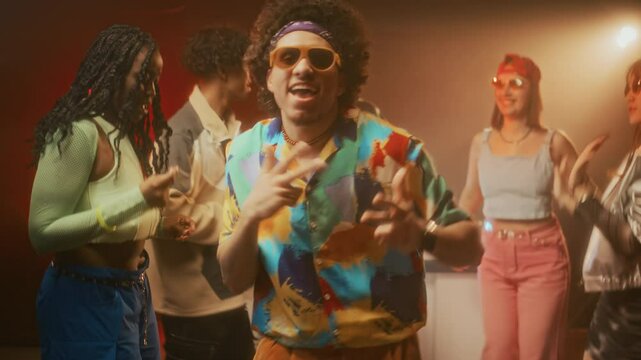 Medium lowkey slowmo of confident and cheeky guy with puffy hair wearing retro hipster clothes and aviator sunglasses dancing confidently to upbeat music and looking at camera at retro party in club