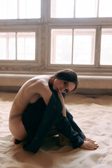 Young sexy woman in an unbuttoned jacket and bare breasts and jeans posing against the wall. Model shooting in an interior studio with sand on the floor.