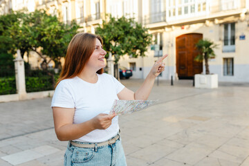 Attractive female tourist is exploring new city. Redhead woman pointing finger something and holding a paper map in old town, Spain or Italy. Traveling Europe