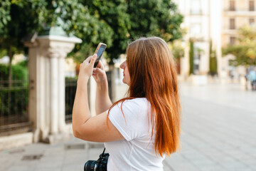 Redhead female traveler in casual dress taking picture of city street with smartphone, travel in Europe. Attractive female tourist is exploring new city. Hight quality photo