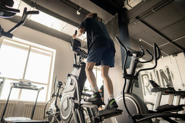 young teenager is working out on an elliptical trainer in a modern gym with large windows. The gym is brightly lit with the sunlight streaming through the windows