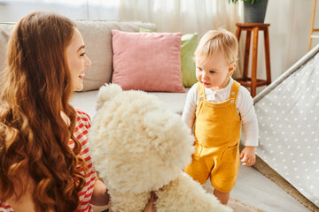 A young mother lovingly holds a teddy bear while her toddler daughter looks on with joy and affection.