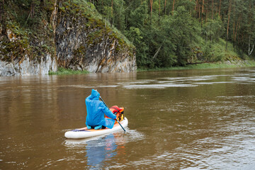 A man in a blue jacket leisurely paddles a canoe along a peaceful river, surrounded by a serene...
