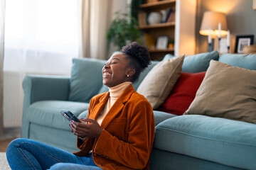 Obraz premium Beautiful black woman feeling very happy after reading a message on her smart phone. She is sitting on the floor in her apartment.