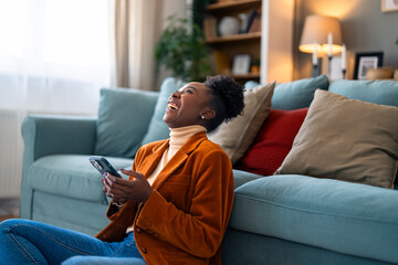 Obraz premium Beautiful black woman feeling very happy after reading a message on her smart phone. She is sitting on the floor in her apartment and reading excited news.