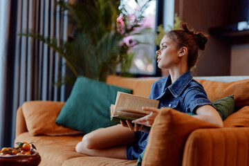 Woman sitting on couch reading book and looking out window in cozy living room setting