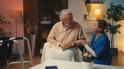 Caring female nurse talking with senior male reading a book, home care services - Powered by Adobe