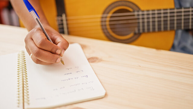 A woman's hand writing lyrics in a notebook on a wooden table with an acoustic guitar in the background, indoors.