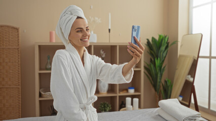 A beautiful young woman with a towel on her head is taking a selfie in a wellness spa room, dressed in a white robe, smiling in a relaxing indoor setting.