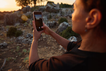 Woman capturing a scenic sunset landscape with her smartphone in an outdoor rocky terrain