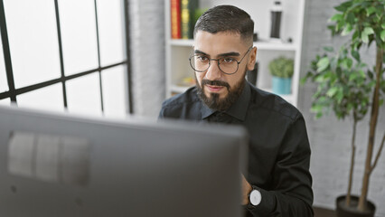 A young adult man with a beard and glasses working focused in a modern indoor office setting.
